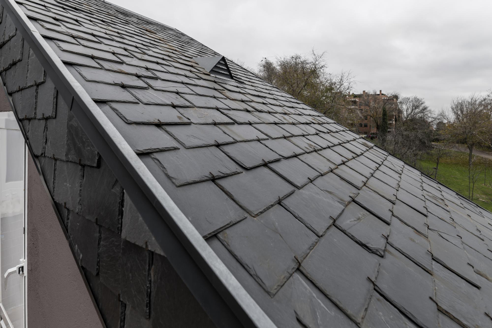 Slate roofing tiles on a roof with a building and trees in the background